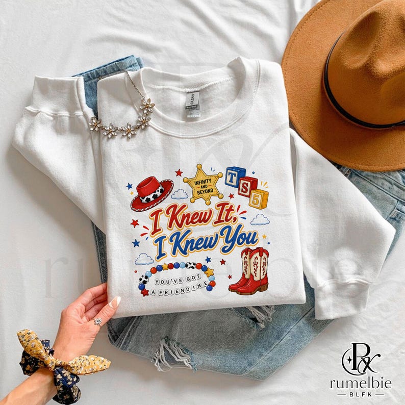 May include: A white long-sleeve sweatshirt featuring a colorful graphic print with a cowboy hat, a sheriff badge labeled Infinity and Beyond, alphabet blocks, red cowboy boots, and a beaded necklace. The text reads I Knew It, I Knew You.