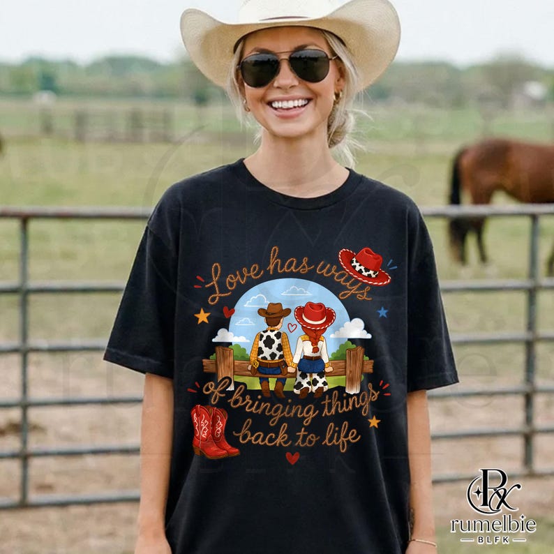 May include: A black t-shirt featuring a graphic of two figures wearing cowboy hats and western attire sitting on a wooden fence. The text reads Love has ways of bringing things back to life. The design includes red cowboy boots and stars.