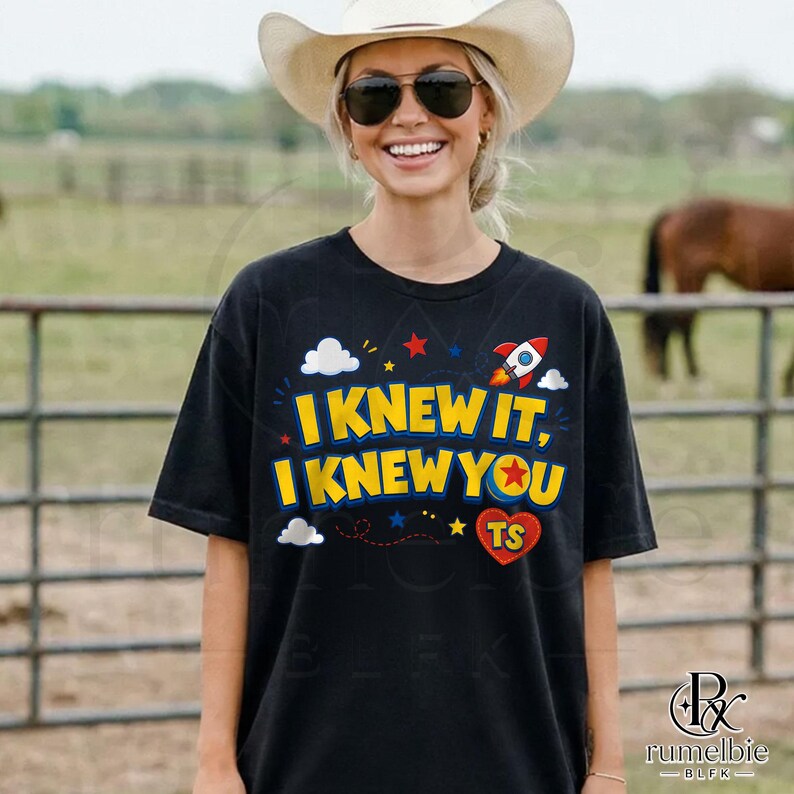 May include: A black short-sleeved t-shirt featuring the yellow text I KNEW IT, I KNEW YOU with a red heart containing the letters TS, a small rocket ship, and white clouds. The design includes small red and blue stars scattered across the front.