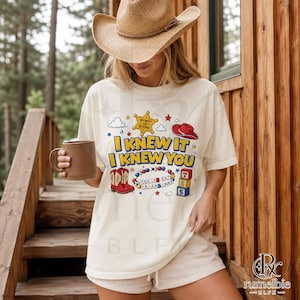 May include: An off-white short-sleeve t-shirt featuring a graphic print with the text I KNEW IT I KNEW YOU, a yellow sheriff badge, a red cowboy hat, red cowboy boots, a beaded bracelet with the text YOU'VE GOT A FRIEND IN ME, and blocks.