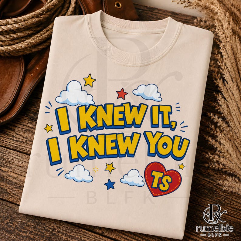 May include: A beige t-shirt featuring the yellow text I KNEW IT, I KNEW YOU with blue outlines. The design includes white clouds, yellow and red stars, and a red heart with yellow TS letters inside, all arranged in a playful, cartoon-style graphic.