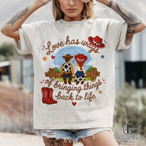 May include: A cream-colored short-sleeve t-shirt featuring a graphic of a cowboy and cowgirl sitting on a wooden fence. The text reads Love has ways of bringing things back to life. The design includes red cowboy boots, hearts, and stars.