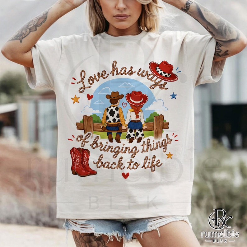 May include: A cream-colored short-sleeve t-shirt featuring a graphic of a cowboy and cowgirl sitting on a wooden fence. The text reads Love has ways of bringing things back to life. The design includes red cowboy boots, hearts, and stars.