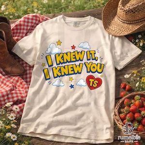 May include: A cream-colored short-sleeve t-shirt featuring the text I KNEW IT, I KNEW YOU in bold yellow letters with blue outlines. The design includes blue and white clouds, small red, yellow, and blue stars, and a red heart with TS inside.