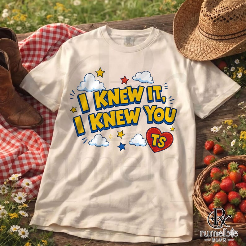 May include: A cream-colored short-sleeve t-shirt featuring the text I KNEW IT, I KNEW YOU in bold yellow letters with blue outlines. The design includes blue and white clouds, small red, yellow, and blue stars, and a red heart with TS inside.