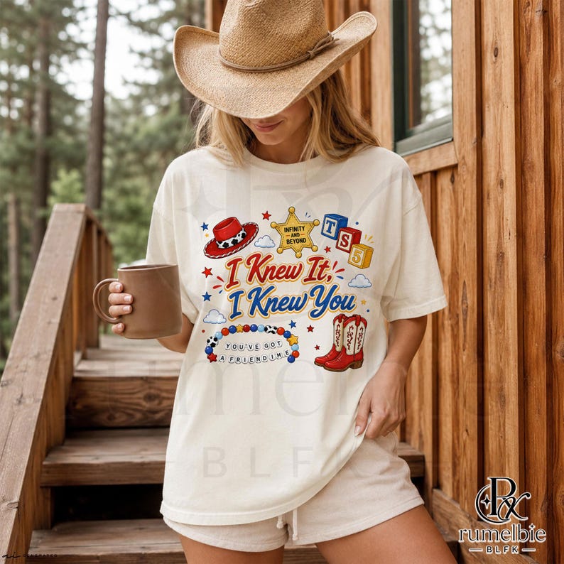 May include: An off-white short-sleeve t-shirt featuring a graphic print with a red cowboy hat, a yellow sheriff star, blue and red alphabet blocks, a beaded bracelet, and red cowboy boots with the text I Knew It, I Knew You, and You've Got A Friend In Me.