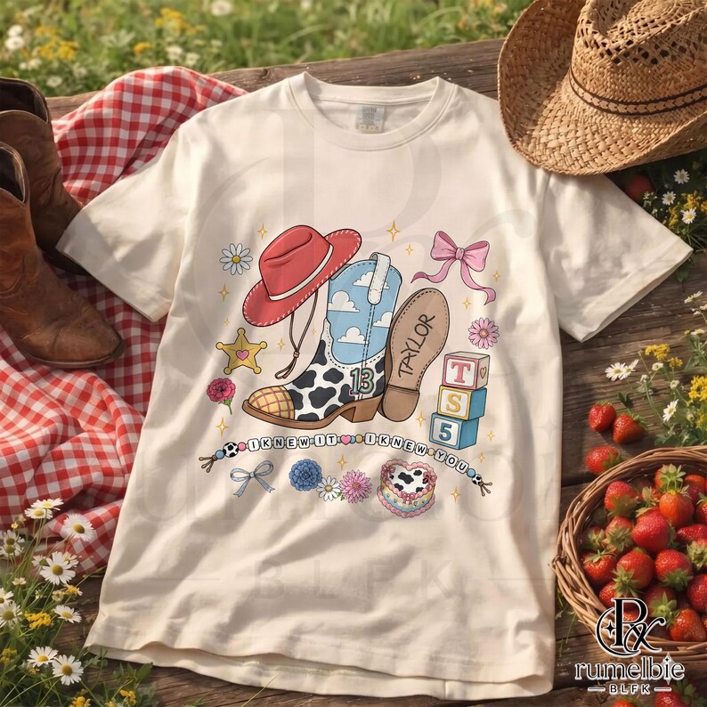 May include: A cream-colored t-shirt featuring a graphic print of a red cowboy hat, a blue and white cow-print cowboy boot with the number 13, a pink bow, and alphabet blocks spelling TS 5. The text I KNEW IT I KNEW YOU is printed below.