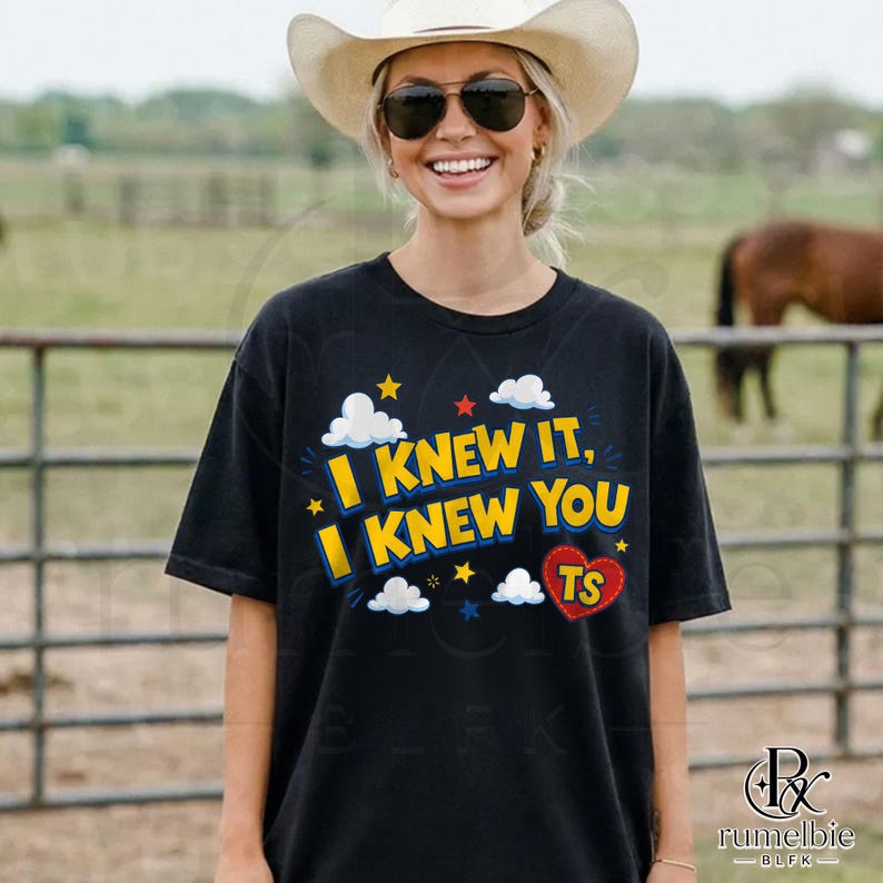May include: A black short-sleeved t-shirt featuring the yellow text "I KNEW IT, I KNEW YOU" with white clouds, small yellow and red stars, and a red heart shape containing the letters "TS" printed on the front of the garment.