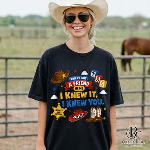 Puede incluir: Una camiseta negra con un estampado gr&aacute;fico que incluye un sombrero de vaquero marr&oacute;n, un sombrero de vaquero rojo y blanco con estampado de vaca y botas de vaquero marrones. El texto dice: You've got a friend in me, I knew it, I knew you.
