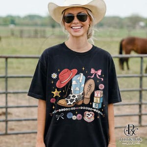 May include: A black short-sleeve t-shirt featuring a graphic print of a red cowboy hat, a blue and white cow-print boot with the number 13, a brown boot sole labeled Taylor, pink bows, flowers, stars, and three stacked blocks labeled T, S, 5.