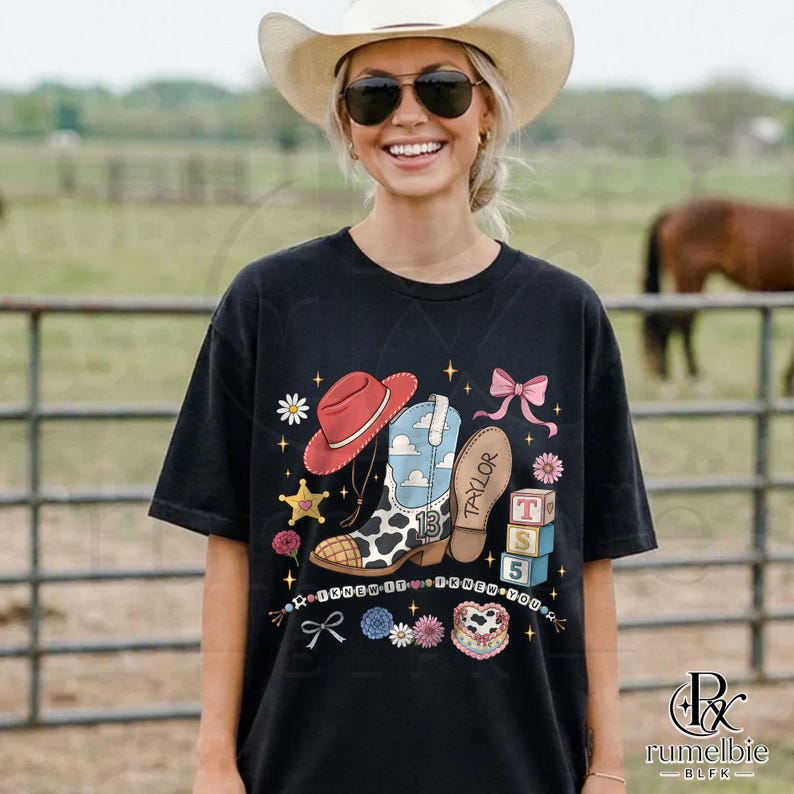 May include: A black short-sleeve t-shirt featuring a graphic print of a red cowboy hat, a blue and white cow-print boot with the number 13, a brown boot sole labeled Taylor, pink bows, flowers, stars, and three stacked blocks labeled T, S, 5.