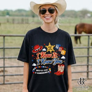 May include: A black t-shirt featuring a graphic print with a red cowboy hat, a gold sheriff star badge with the text Infinity and Beyond, blue and red alphabet blocks, a colorful beaded bracelet, and red cowboy boots with the text I Knew It I Knew You.