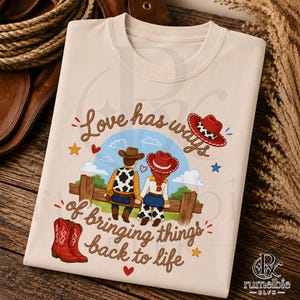 May include: A cream-colored t-shirt featuring a graphic of two figures in cowboy attire sitting on a wooden fence. The text reads, Love has ways of bringing things back to life. The design includes red cowboy boots, stars, and hearts.