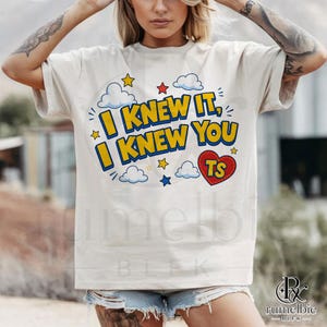 May include: A cream-colored short-sleeve t-shirt featuring a graphic print with the text I KNEW IT, I KNEW YOU in bold yellow letters with blue outlines. The design includes blue and white clouds, small yellow and red stars, and a red heart with TS.