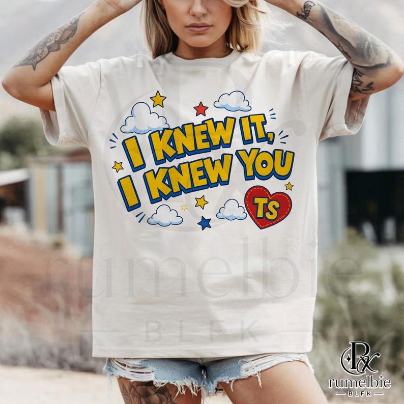 May include: A cream-colored short-sleeve t-shirt featuring a graphic print with the text I KNEW IT, I KNEW YOU in bold yellow letters with blue outlines. The design includes blue and white clouds, small yellow and red stars, and a red heart with TS.