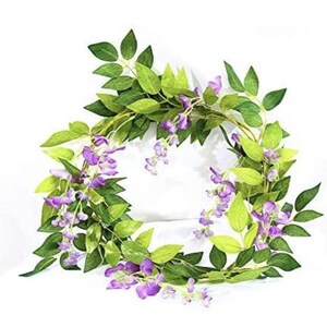 May include: A wreath of artificial wisteria vines with green leaves and clusters of purple flowers. The wreath is arranged in a circular shape, suitable for home decor or seasonal displays.