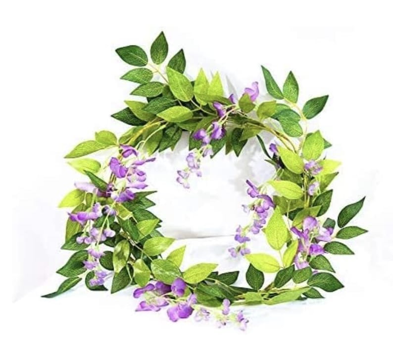 May include: A wreath of artificial wisteria vines with green leaves and clusters of purple flowers. The wreath is arranged in a circular shape, suitable for home decor or seasonal displays.