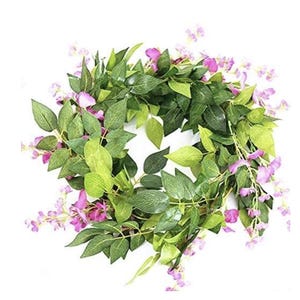 May include: A circular wreath composed of green leaves and small purple flowers. The leaves vary in shades of green, creating depth and texture. The wreath is designed for decorative purposes, suitable for seasonal or year-round display.