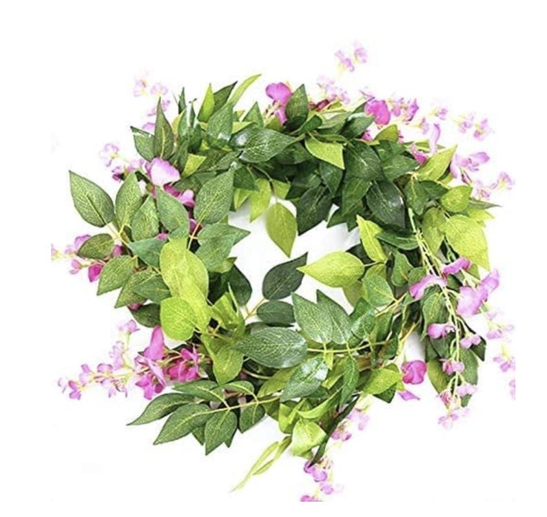 May include: A circular wreath composed of green leaves and small purple flowers. The leaves vary in shades of green, creating depth and texture. The wreath is designed for decorative purposes, suitable for seasonal or year-round display.
