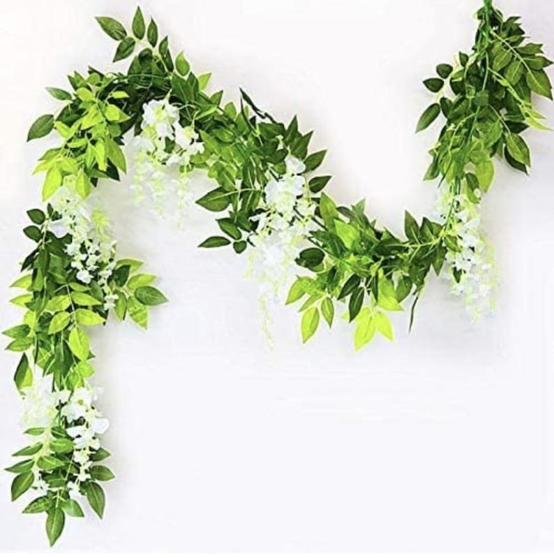 May include: Artificial wisteria garland with white flowers and green leaves. The garland is arranged in a flowing, decorative pattern, perfect for adding a touch of nature to any space. The leaves have varying shades of green.