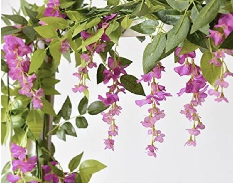May include: Artificial wisteria garland with cascading purple flowers and green leaves. The flowers are a vibrant shade of purple, and the leaves are a deep green. The garland is designed for decorative purposes.