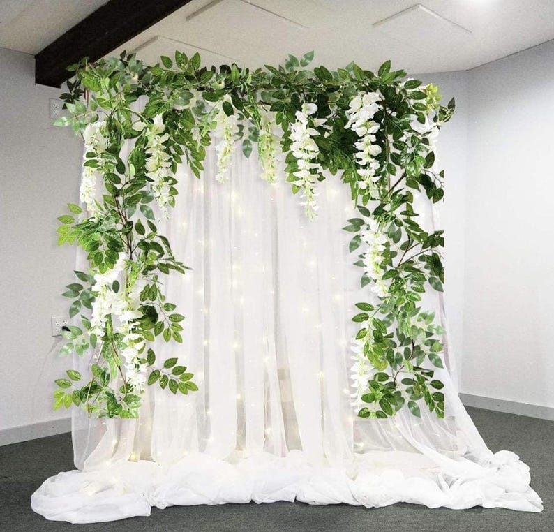 May include: A decorative backdrop featuring white sheer fabric, adorned with cascading white wisteria and lush green foliage. Small lights are visible behind the fabric, creating a soft glow. Ideal for weddings or special events.