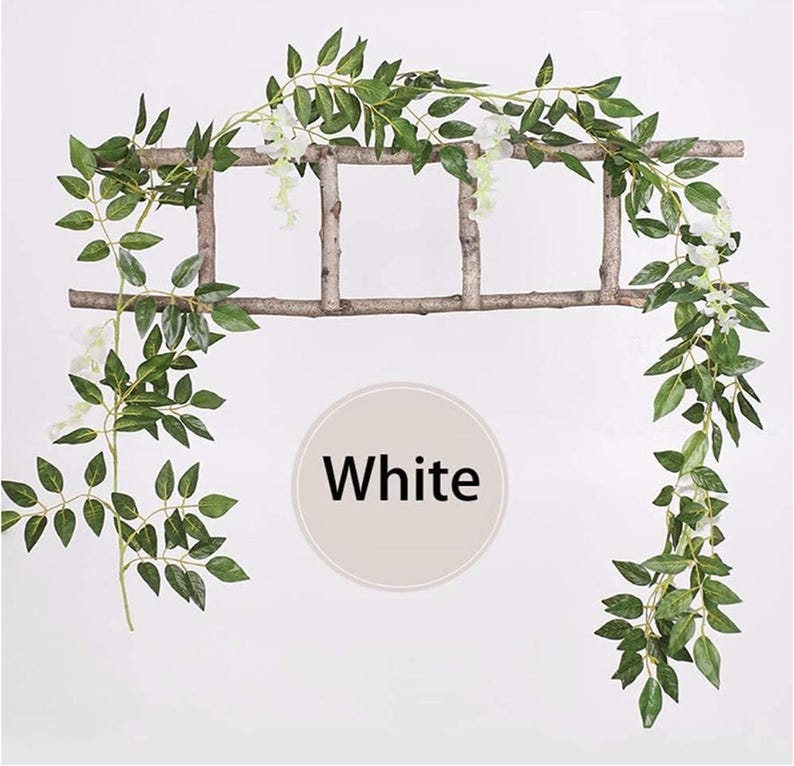 May include: A decorative garland featuring green leaves and white flowers, draped over a rustic wooden ladder. The word "White" is displayed in a white circle. Ideal for home decor or event styling.