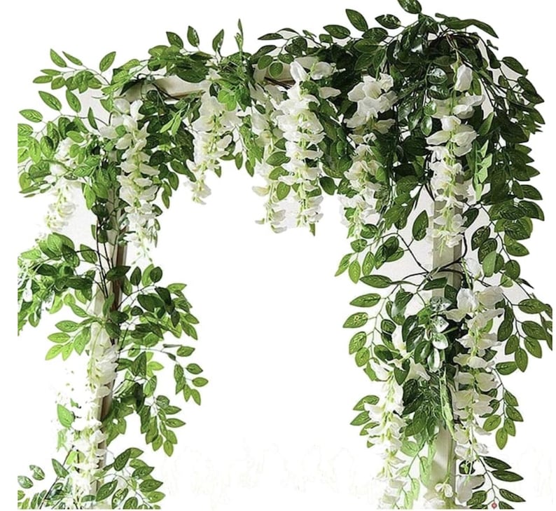 May include: Artificial wisteria garland with white flowers and green leaves. The garland is arranged in an arch shape, suitable for weddings or other decorative purposes. The flowers are clustered in long, cascading strands.