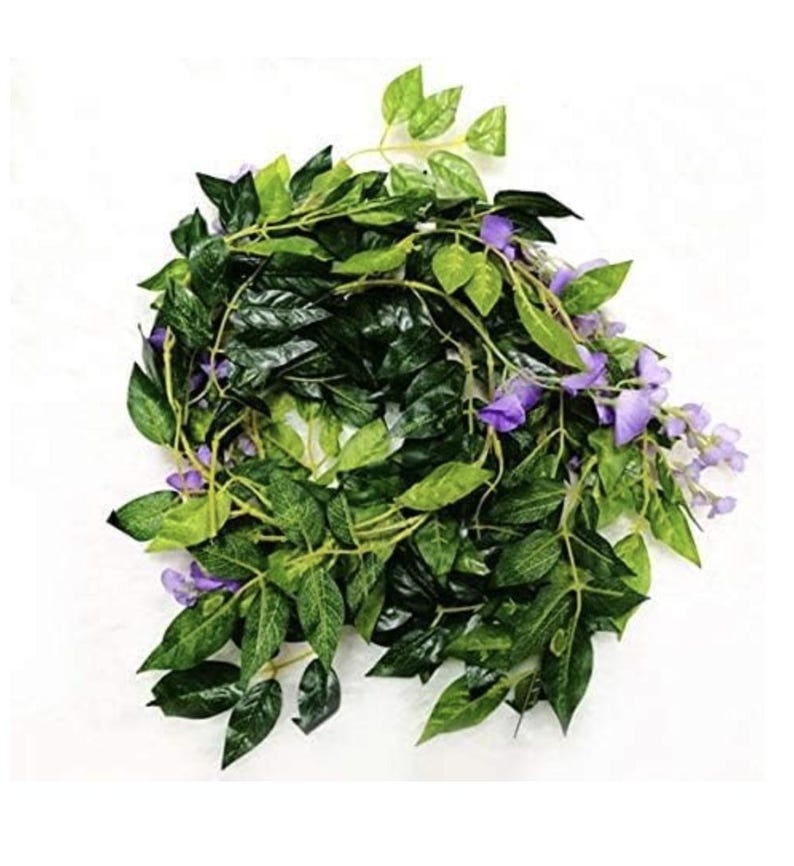 May include: A garland of artificial greenery with dark green leaves and small clusters of purple flowers. The leaves vary in shades of green, creating a realistic appearance. The garland is designed for decorative purposes.