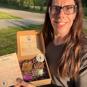 May include: A person holding an open cardboard box containing two mineral specimens, a purple keychain, and a round sticker. The box has text that reads "Welcome to your next station Trailblazer". The box also contains a Field Notes card.
