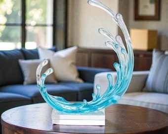 May include: A decorative sculpture of a stylized water wave in shades of clear and blue. The wave is mounted on a white rectangular base and sits on a dark wooden table. The sculpture is in a living room setting.