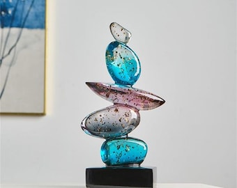May include: A decorative sculpture featuring stacked, oval-shaped glass stones in shades of blue, pink, and clear. The stones have gold flecks and sit atop a black rectangular base. The sculpture is a modern home decor accent.