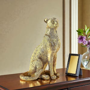 May include: A gold cheetah sculpture with a textured finish, in a seated position. The statue has dark spots and is placed on a dark wooden surface. A small black framed picture and a vase with flowers are in the background.