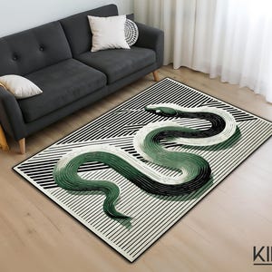 Puede incluir: Una alfombra rectangular con un fondo a rayas blancas y negras y un gran diseño de serpiente verde, blanca y negra. La alfombra está sobre un suelo de madera, con un sofá gris y cojines en el fondo. La marca "KIRAC" está en la esquina inferior derecha.