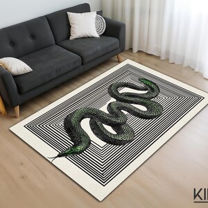 Könnte beinhalten: Rechteckiger Teppich mit schwarz-weißem geometrischen Rand. Der Teppich zeigt eine detaillierte Illustration einer grünen Schlange, die sich in der Mitte windet. Der Teppich liegt auf einem Holzboden. Die Marke "KIRAC" ist sichtbar.