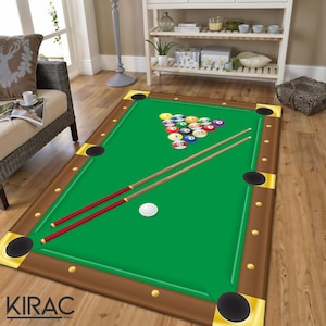 May include: A rectangular green and brown area rug designed to resemble a pool table. The rug features a green playing surface, brown borders, and gold corner accents. A white cue ball and a rack of numbered billiard balls are visible, along with two pool cues.