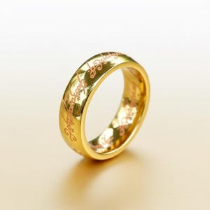 May include: A gold ring with engraved script. The ring has a polished finish and a rounded shape. The engraved text is in a contrasting colour. The ring is set against a neutral background.