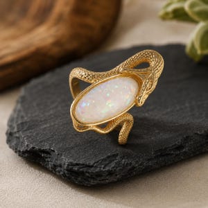 Könnte beinhalten: Ein goldfarbener Ring mit einem strukturierten Schlangendesign, das sich um einen ovalen, weißen Opalstein windet. Der Stein zeigt irisierende Farbreflexe, und das Metallband ist mit Schuppen entlang des Schlangenkörpers und des Kopfes versehen.