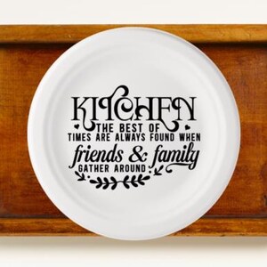 May include: A white plate with black text that reads "KITCHEN THE BEST OF TIMES ARE ALWAYS FOUND WHEN friends & family GATHER AROUND" sits on a wooden tray with metal handles. The tray is a warm brown color.