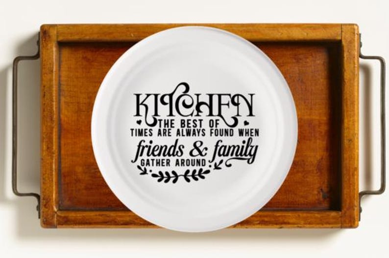 May include: A white plate with black text that reads "KITCHEN THE BEST OF TIMES ARE ALWAYS FOUND WHEN friends & family GATHER AROUND" sits on a wooden tray with metal handles. The tray is a warm brown color.