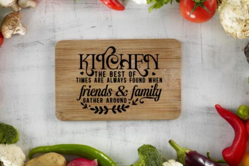 May include: A rectangular wooden cutting board with the word "KITCHEN" in a decorative font. The text "THE BEST OF TIMES ARE ALWAYS FOUND WHEN friends & family GATHER AROUND" is also printed on the board. Vegetables surround the board.
