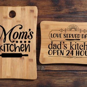 May include: Two wooden cutting boards with black text. One says "Mom's kitchen" with a rolling pin graphic. The other says "Love served daily dad's kitchen OPEN 24 HOURS" with a sun graphic.