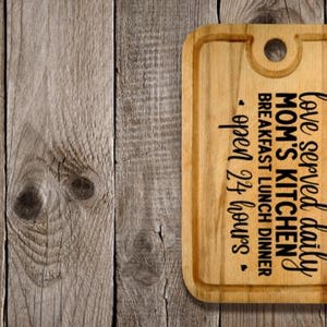 May include: A rectangular wooden cutting board with rounded corners and a handle hole. The board features the text "love served daily, Mom's Kitchen, breakfast lunch dinner, open 24 hours" in black script.