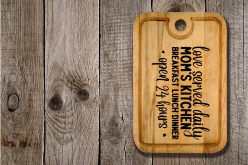 May include: A rectangular wooden cutting board with rounded corners and a handle hole. The board features the text "love served daily, Mom's Kitchen, breakfast lunch dinner, open 24 hours" in black script.