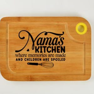 May include: A rectangular bamboo cutting board with the words "Nama's KITCHEN" in a black script font. Below the text is the phrase "where memories are made AND CHILDREN ARE SPOILED" and a whisk illustration. The board has a yellow hole.