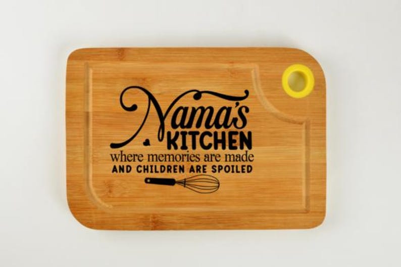 May include: A rectangular bamboo cutting board with the words "Nama's KITCHEN" in a black script font. Below the text is the phrase "where memories are made AND CHILDREN ARE SPOILED" and a whisk illustration. The board has a yellow hole.
