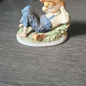 May include: A porcelain figurine of a young person in blue overalls and a straw hat, reclining on a bed of green grass. The figurine has a light skin tone and is resting with their eyes closed. A small, broken pot sits nearby.