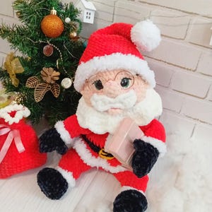 Pode incluir: Boneca de Papai Noel de pel&uacute;cia feita &agrave; m&atilde;o, vestida com um traje vermelho com acabamentos brancos, luvas e botas pretas e um chap&eacute;u a condizer com um pompom branco. A boneca segura um pequeno presente rosa e usa &oacute;culos redondos. Uma pequena &aacute;rvore de Natal e um saco de presente vermelho est&atilde;o no fundo.