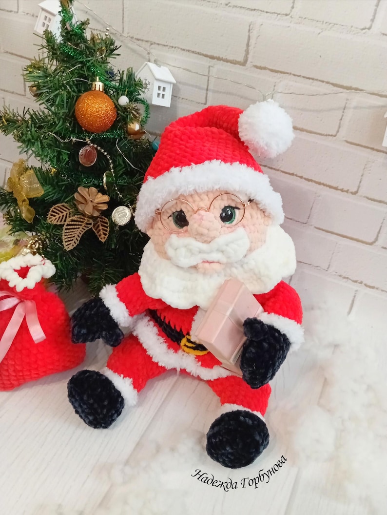 Pode incluir: Boneca de Papai Noel de pel&uacute;cia feita &agrave; m&atilde;o, vestida com um traje vermelho com acabamentos brancos, luvas e botas pretas e um chap&eacute;u a condizer com um pompom branco. A boneca segura um pequeno presente rosa e usa &oacute;culos redondos. Uma pequena &aacute;rvore de Natal e um saco de presente vermelho est&atilde;o no fundo.