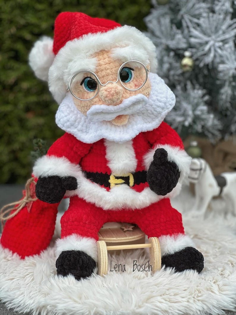 Pode incluir: Boneca de croch&ecirc; feita &agrave; m&atilde;o do Papai Noel, vestindo um terno vermelho com acabamento branco, um chap&eacute;u combinando e &oacute;culos redondos. A boneca tem barba e bigode brancos, luvas pretas e est&aacute; segurando um saco vermelho. O nome "Lena Bosch" est&aacute; na base.